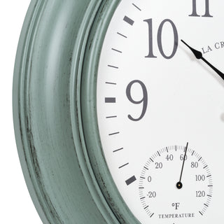 Close-up of a round wall clock showing a distressed sage green frame, white dial, black numerals, and a small temperature gauge at the bottom right.
