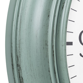Close-up of a distressed wall clock showing a muted sage green painted frame with dark weathered wood showing through, next to a white clock face with black markings.
