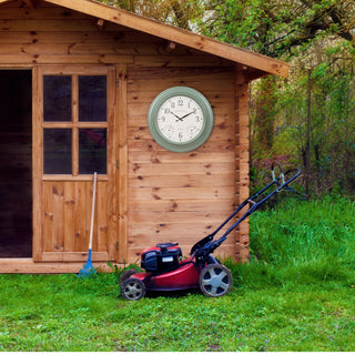 A red and black push lawn mower sits on green grass in front of a wooden cabin, with a round wall clock above it and a blue-handled broom leaning against the door.