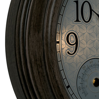 Close-up of a decorative wall clock showing a dark wood frame, a patterned silver dial, black Arabic numerals, and a small gold-toned center detail against a white background.