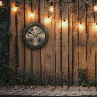 A round vintage-style wall clock with a dark green frame and distressed beige face, mounted on a wooden fence outdoors. Warm string lights glow in the background above a stone patio.