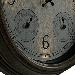 Close-up of a decorative wall clock face with two analog dials, a large black numeral 6, and an etched leaf pattern on a light gray dial.
