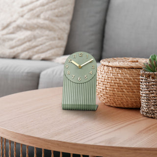 Lifestyle image of the 437-3015LS Grooves Tabletop Clock shown on a wood side table next to a couch. The clock features a molded light sage green L-shape design with textured grooves, an open face dial, step motion quartz movement, and brass hands and numbers.