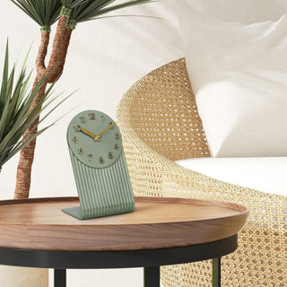 Lifestyle image of the 437-3015LS Grooves Tabletop Clock shown on a wood side and metal table next to a cream and rattan chair. The clock features a light sage green molded L-shape design with textured grooves, an open face dial, step motion quartz movement, and brass hands and numbers.