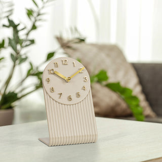 Right-facing lifestyle image of the 437-3015SW Grooves Tabletop Clock shown on a neutral countertop with plants and a couch in the background. The clock features a soft white molded L-shape design with textured grooves, an open face dial, step motion quartz movement, and brass hands and numbers.