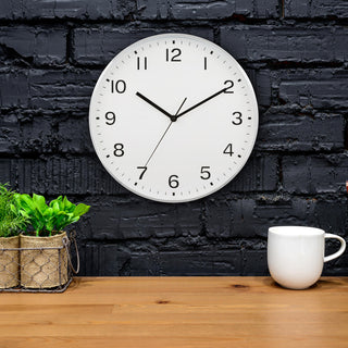 Lifestyle image of the 46201 13-inch Brushed Frame Wall Clock shown hanging on a dark gray brick wall above a wood table with plants and a coffee cup. The clock features a white dial with black numbers and hands in a brushed silver case.