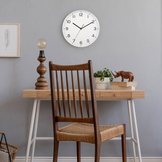 Lifestyle image of the 46201 13-inch Brushed Frame Wall Clock shown hanging on a grey wall avouve a Scandinavian style desk with a wood lamp, books, and a wood carving of a rhinocerous. The clock features a white dial with black numbers and hands in a brushed silver case.