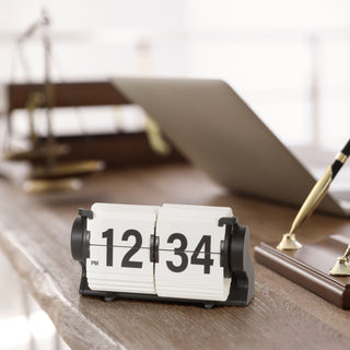 Lifestyle image of the 536-527W Rollins Mini Mechanical Flip Clock – White, shown sitting on a dark wood live-edge desk next to a wood base pen set in gold and black with a computer in the background. The clock features white hour and minute flip cards with black numbers in a black case.