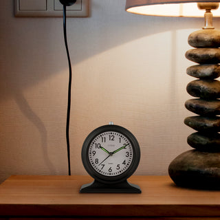 617-3014 A round black analog desk clock with a white face and green hands sits on a wooden table next to a lit lamp and a stack of stacked stones.