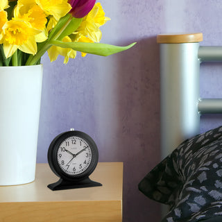 617-3014 A black analog desk clock with a white face and black base sits on a light wood bedside table next to a white vase of yellow daffodils and a bed with a patterned pillow.