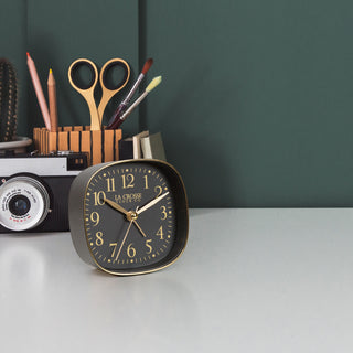Light gray desk with a La Crosse Clock Co. retro black and gold alarm clock, scissors, pens, and a camera with dark green wall.