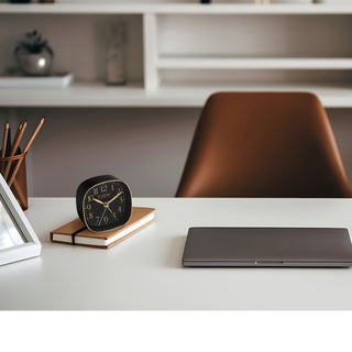 A modern desk setup in a transitional décor style with a black and gold tabletop alarm clock on a notebook with a laptop, pencil cup and frame.