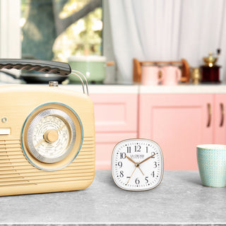 A vintage radio next to a retro style white and gold table top alarm clock, and a cup on a kitchen countertop.