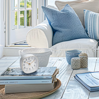 Cozy coastal décor living room  with a La Crosse Clock Co. white and gold alarm clock sitting on top of books on a weathered wood coffee table, a white couch with blue cushions, and decorative items on a table.