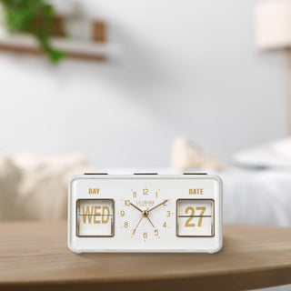 Lifestyle image of the 636-379W Daily Flip Tabletop Clock with Calendar – White, shown sitting on a light wood table. The retro rectangle design clock features an analog clock with brass numbers, hour, minute, and second hands, and a day and date calendar with manual flip cards with brass text.