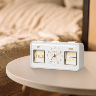 Lifestyle image of the 636-379W Daily Flip Tabletop Clock with Calendar – White, shown sitting on a neutral side table with faux fur tan bedding in the background. The retro rectangle design clock features an analog clock with brass numbers, hour, minute, and second hands, and a day and date calendar with manual flip cards with brass text.