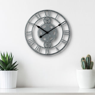 Lifestyle image of the 83186-BHG 14-inch Metal Gear Clock shown hanging on a white wall above a white shelf with white potted plants. The clock features a distressed gray metal skeleton design with gears, Roman numerals, and black metal hands.