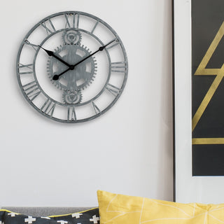 Lifestyle image of the 83186-BHG 14-inch Metal Gear Clock shown hanging on a white wall with black and gold accents above a gray couch. The clock features a distressed gray metal skeleton design with gears, Roman numerals, and black metal hands.