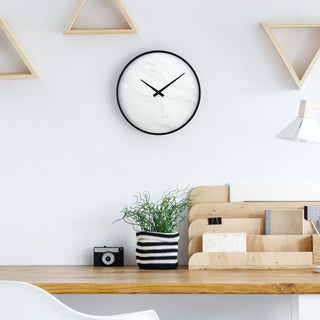 Lifestyle image of the 83216-BHG 14-inch Faux Marble Wall Clock shown hanging on a white wall above a medium wood tone desk with a white chair, wood desk organizer, a plant in a black and white striped knitted pot, and geometric wall art. The Clock features a thin black frame, a faux-marble face, no numbers or markings, and modern black hands.