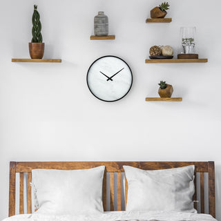 Lifestyle image of the 83216-BHG 14-inch Faux Marble Wall Clock shown hanging on a white wall above a slotted wood bedframe with white bedding and wood shelves on the wall holding organic vases, plants, and decorations. The Clock features a thin black frame, a faux-marble face, no numbers or markings, and modern black hands.