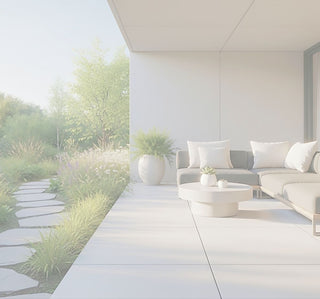 Modern outdoor patio seating area with a dark gray sectional sofa, white throw pillows, a round white coffee table, and a large potted plant, set beside a landscaped garden with a stone path and tall grasses.