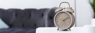 A classic twin-bell alarm clock with a beige body and gold accents, sitting on a white table in a modern living room setting with a blurred gray sofa in the background.
