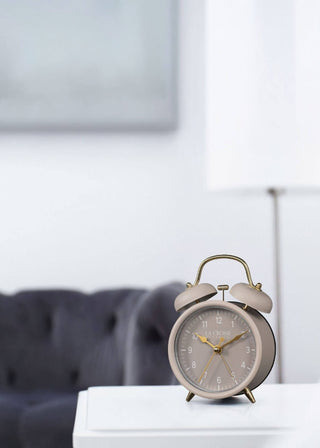 A classic twin-bell alarm clock with a beige body and gold accents, sitting on a white table in a modern living room setting with a blurred gray sofa in the background.