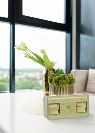 A retro-style dual-date alarm clock in sage green sits on a white table in a modern living room setting, styled with a gold vase of tulips and succulents in the background.