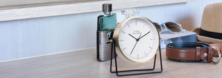 A round analog desk clock with a gold-toned frame and black stand, styled on a light wood surface alongside a green bottle, sunglasses, a brown leather belt, and a tan hat.