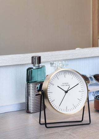 A round analog desk clock with a gold-toned frame and black stand, styled on a light wood surface alongside a green bottle, sunglasses, and a brown leather belt.