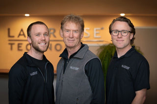 Three men standing together indoors wearing matching black and gray branded apparel, with a large 'La Crosse Technology' sign visible in the background.