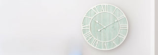 A round wall clock with a distressed light green wood-grain face, white Roman numerals, and white hands, mounted on a plain white wall.