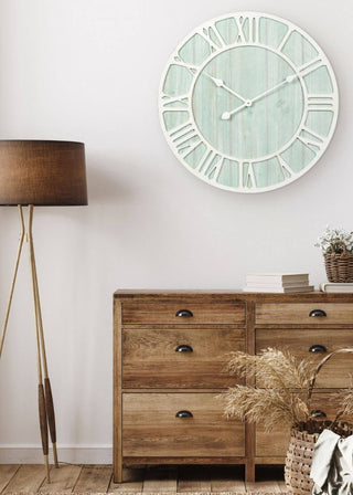 A large round wall clock with a distressed mint green face and white Roman numerals, mounted on a white wall above a rustic wooden dresser in a styled room setting.