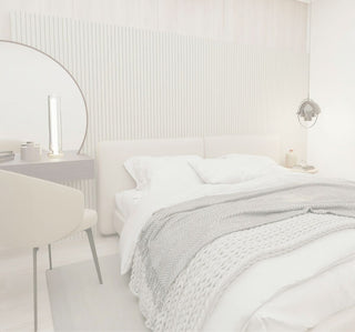 A modern bedroom featuring a bed dressed in white linens and a gray textured throw blanket, set against a light wood slatted accent wall. A matching beige upholstered chair and a dark vanity table with a large oval mirror are visible on the left.