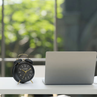 Lifestyle image of the W72449 Metal Twin Bell Alarm Clock – Black, sitting on a desk next to a laptop with a window overlooking a green yard in the background. The clock features a black dial with white numbers, gold hands and accents, in a black metal case.