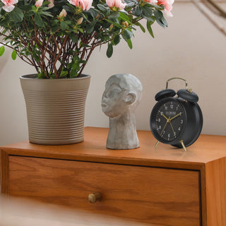 Lifestyle image of the W72449 Metal Twin Bell Alarm Clock – Black, sitting on a wood side table in a foyer with a bust, and a plant. The clock features a black dial with white numbers, gold hands and accents, in a black metal case.