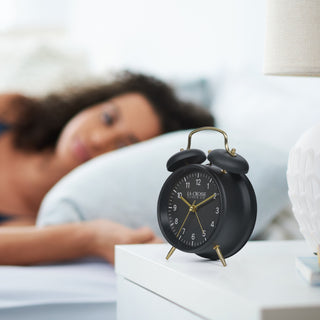 Lifestyle image of the W72449 Metal Twin Bell Alarm Clock – Black, sitting on a white night stand with a woman sleeping in the background The clock features a black dial with white numbers, gold hands and accents, in a black metal case.
